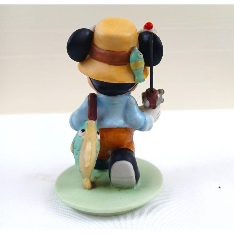 May include: A figurine of Mickey Mouse dressed as a fisherman, viewed from the back. He wears a yellow hat with a brown band, a light blue shirt, orange pants, and black shoes. He holds a fishing rod and a fish. The figurine stands on a green base.