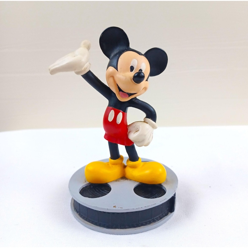 May include: A Mickey Mouse figurine in a cheerful pose, wearing red shorts, yellow shoes, and white gloves. The black-eared character stands atop a gray film reel base, with one arm raised. The figurine has a smiling expression.