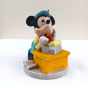 May include: A figurine of Mickey Mouse, wearing a blue beret, yellow shirt, and red bow tie, is depicted working at a yellow desk. The desk has a gray printing press and a roll of yellow paper. The figurine is on a gray base.
