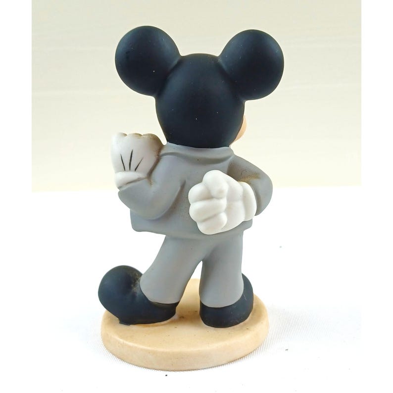 Pode incluir: Uma estatueta do Mickey Mouse vista por tr&aacute;s, vestindo um terno cinza e luvas brancas. A figura tem orelhas e sapatos pretos e est&aacute; sobre uma base circular bege. Um item de colecionador cl&aacute;ssico da Disney.