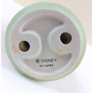 May include: Bottom view of a ceramic figurine base with a light green rim. The base is white with two holes and the Disney logo and "Sri Lanka" printed on it.