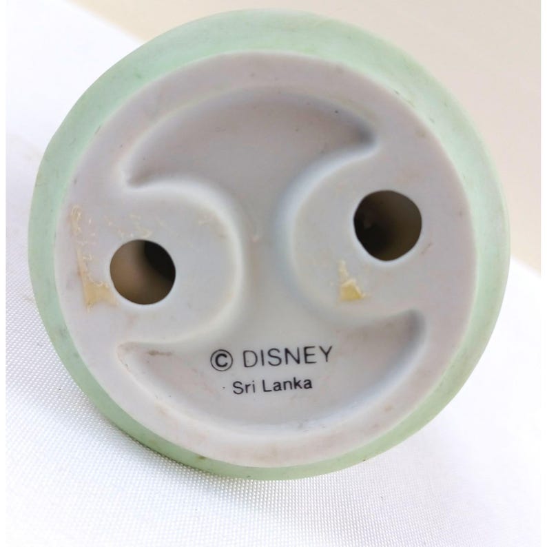 May include: Bottom view of a ceramic figurine base with a light green rim. The base is white with two holes and the Disney logo and "Sri Lanka" printed on it.