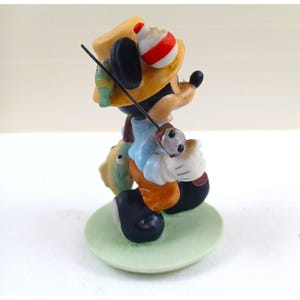 May include: A figurine of Mickey Mouse fishing. Mickey is wearing a yellow hat, blue shirt, and orange pants. He is holding a fishing rod and a fish. The figurine is on a green circular base.