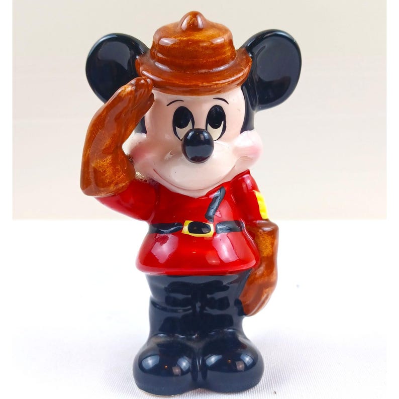 May include: A ceramic figurine of Mickey Mouse in a red uniform, black pants, and a brown hat, saluting with his right hand. The figurine has a black nose, ears, and a yellow patch on the shoulder.