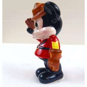 May include: A ceramic figurine of Mickey Mouse in a red jacket, black pants, and a brown hat. Mickey is holding his hand up to his forehead, as if saluting or looking out at something. The figurine has a glossy finish.