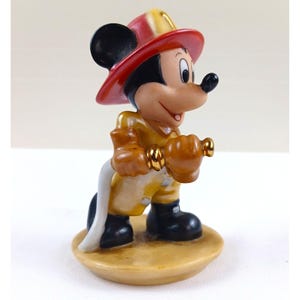 May include: A figurine of Mickey Mouse dressed as a firefighter. Mickey wears a red and yellow fire helmet, a yellow uniform, and black boots. He holds a gold-colored nozzle and a white hose. The figurine is on a tan base.