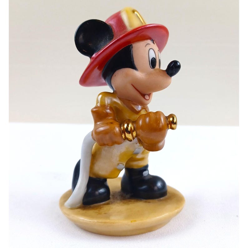 May include: A figurine of Mickey Mouse dressed as a firefighter. Mickey wears a red and yellow fire helmet, a yellow uniform, and black boots. He holds a gold-colored nozzle and a white hose. The figurine is on a tan base.