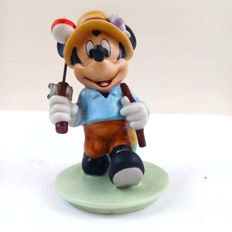May include: A ceramic figurine of Mickey Mouse dressed as a fisherman. Mickey wears a yellow hat, blue shirt, and brown pants. He holds a fishing rod with a red and white bobber and a brown walking stick. The figurine stands on a light green base.