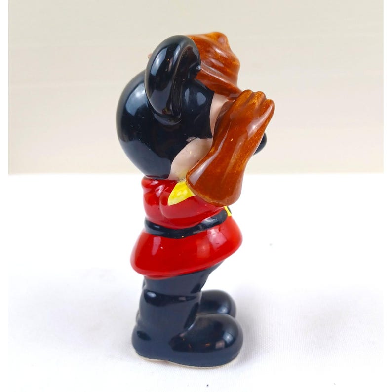 May include: A vintage figurine of a cartoon character, featuring a black head with large ears, a red jacket, and black boots. The character is holding something up to its face with brown gloves. The figurine has a yellow collar.