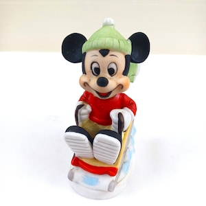 May include: A ceramic Mickey Mouse figurine, wearing a red top and green hat, seated on a yellow and blue sled. The figurine has black ears, white gloves and shoes. A collectible item.