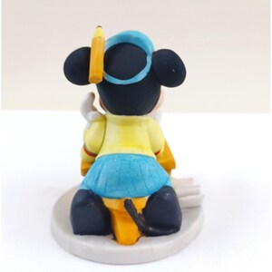 May include: A figurine of a cartoon mouse wearing a blue hat and skirt, and a yellow shirt. A yellow pencil is placed on the mouse's head. The mouse is sitting on a white base.