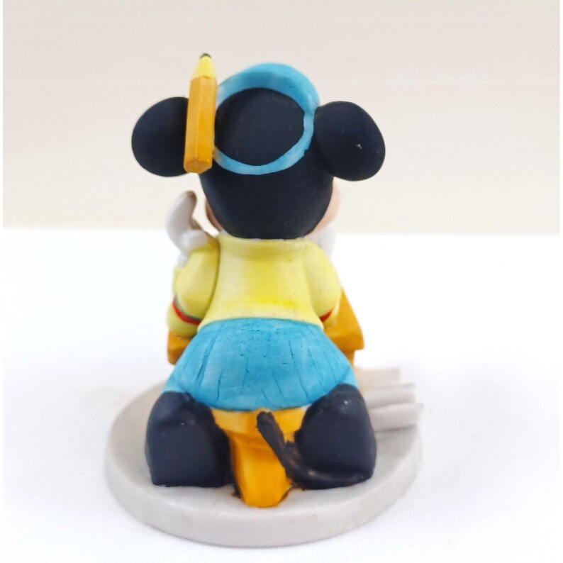 May include: A figurine of a cartoon mouse wearing a blue hat and skirt, and a yellow shirt. A yellow pencil is placed on the mouse's head. The mouse is sitting on a white base.