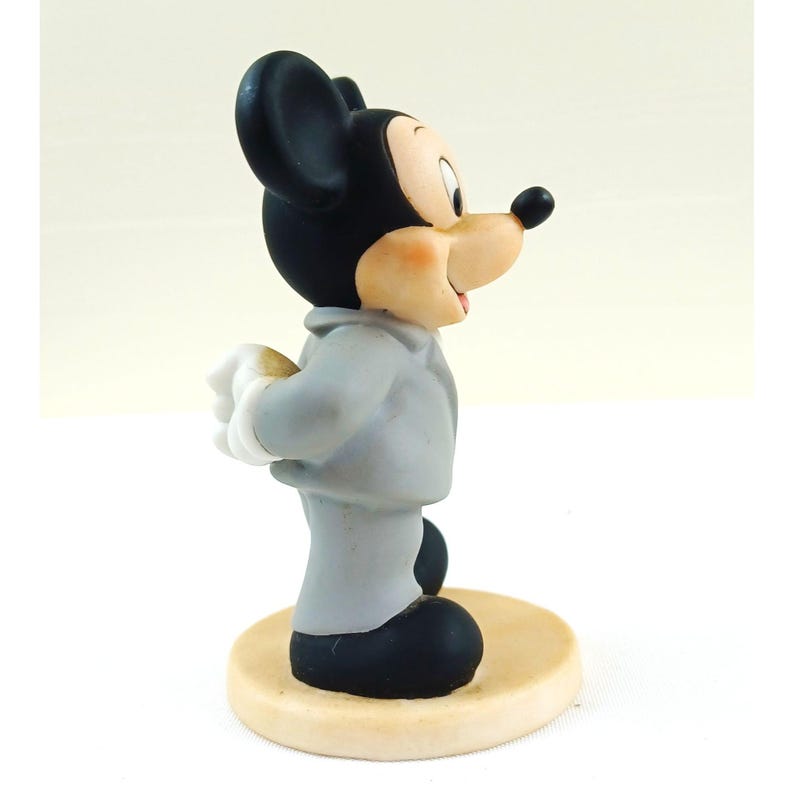 Pode incluir: Uma estatueta do Mickey Mouse com um fato cinzento, sapatos pretos e orelhas pretas. A figura est&aacute; sobre uma base circular bege, com luvas brancas e uma express&atilde;o sorridente. Um item de colec&ccedil;&atilde;o.