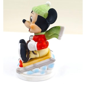 May include: A ceramic figurine of Mickey Mouse skiing. Mickey is wearing a green hat, red jacket, yellow trousers, and white gloves. He has a green scarf and is on a sled with skis. The base is white with blue accents.