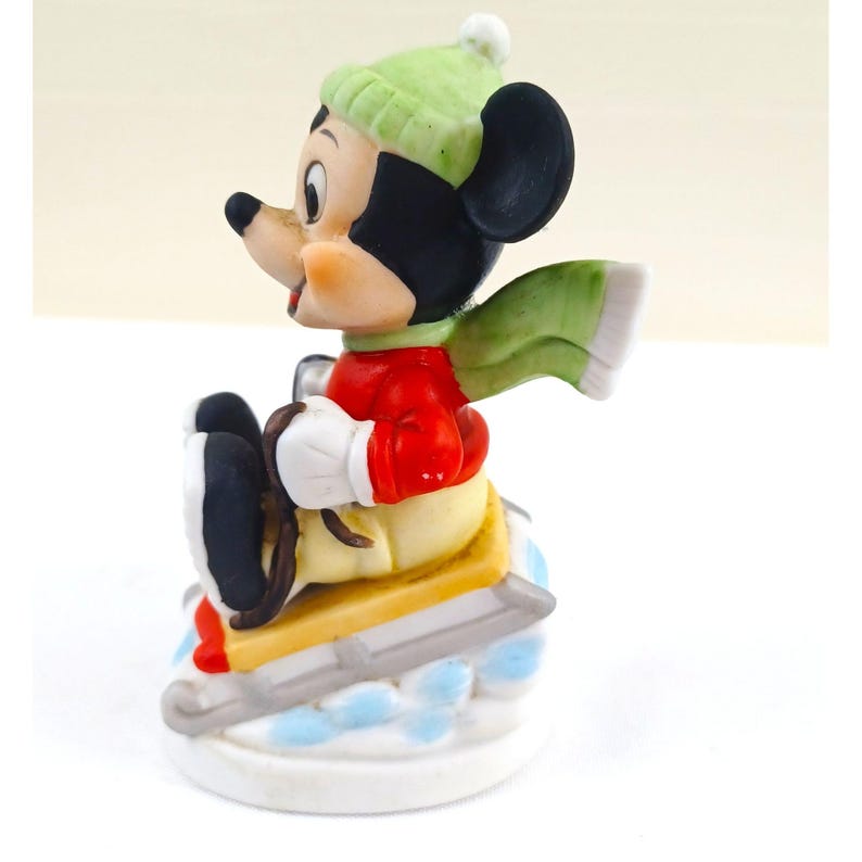 May include: A ceramic figurine of Mickey Mouse skiing. Mickey is wearing a green hat, red jacket, yellow trousers, and white gloves. He has a green scarf and is on a sled with skis. The base is white with blue accents.