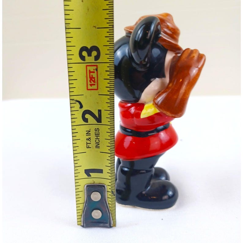 May include: A vintage figurine of a cartoon character, approximately 2.5 inches tall. The figurine is wearing a red jacket, black pants, and black boots. The character has black ears, brown hair, and brown gloves. The figurine is standing on a white surface.
