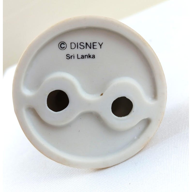 Pode incluir: Uma vista de perto da base de uma estatueta de cer&acirc;mica branca. A base tem dois orif&iacute;cios circulares e um design ondulado. O texto "&copy; DISNEY Sri Lanka" est&aacute; impresso na base.
