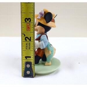 May include: A figurine of a cartoon character dressed as a fisherman, wearing a yellow hat and holding a fish. The figurine is approximately 2 inches (5 cm) tall and stands on a green base.