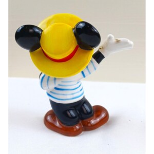 May include: A ceramic figurine of a cartoon character wearing a yellow hat with a red band, black ears, and a blue and white striped shirt. The character is wearing black pants and brown shoes, with one white-gloved hand raised.
