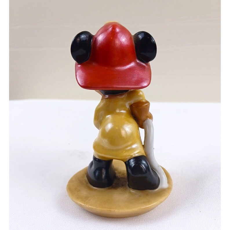 May include: A vintage figurine of a cartoon character dressed as a firefighter. The figure wears a red helmet, yellow coat, and black boots. It holds a white hose and stands on a tan base. The figurine is a collectible item.