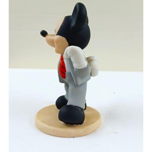 Pode incluir: Uma estatueta do Mickey Mouse em um terno cinza com uma gravata vermelha, luvas brancas e sapatos pretos. A figura est&aacute; em uma base circular bege. O personagem de desenho animado est&aacute; voltado para a esquerda.