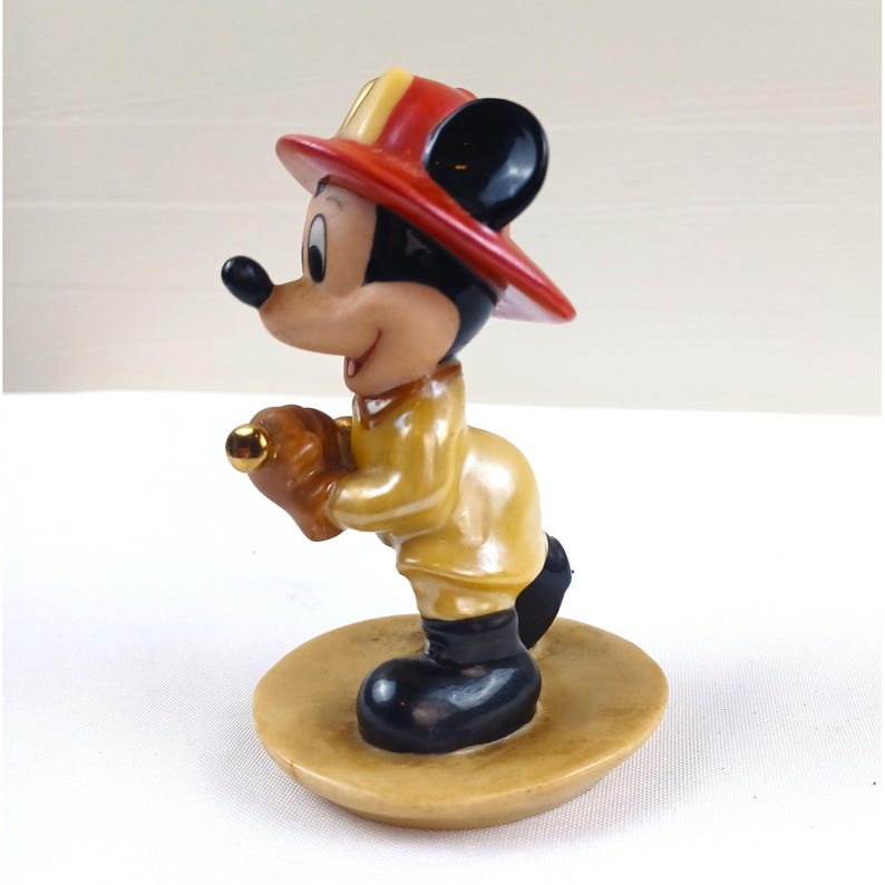 May include: A figurine of Mickey Mouse dressed as a firefighter. Mickey wears a yellow coat, black boots, and a red and yellow firefighter's hat. He is holding a brown glove with a gold accent. The figurine is on a tan base.