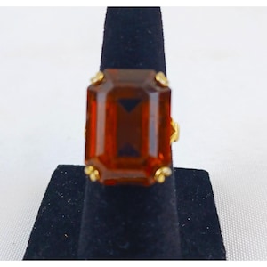 May include: A gold-toned ring featuring a large, rectangular, amber-colored gemstone. The gemstone has a dark, geometric design in the center. The ring is displayed on a black stand against a white background.