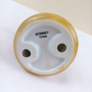 May include: Bottom view of a ceramic figurine base. The base is white with two holes and a curved design. The top edge is tan. The text "&copy; DISNEY CHINA" is printed on the base.