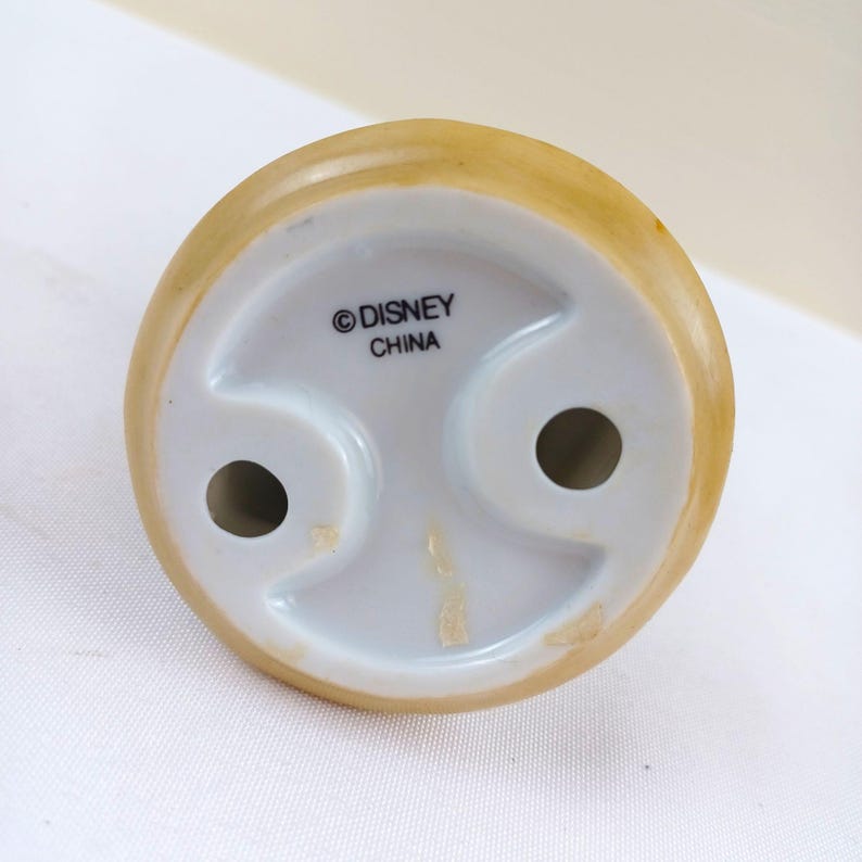 May include: Bottom view of a ceramic figurine base. The base is white with two holes and a curved design. The top edge is tan. The text "&copy; DISNEY CHINA" is printed on the base.