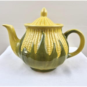 May include: A ceramic teapot with a corn-on-the-cob design. The teapot features a yellow lid and spout, a green base, and a yellow and green corn pattern around the body. The handle is a curved loop.