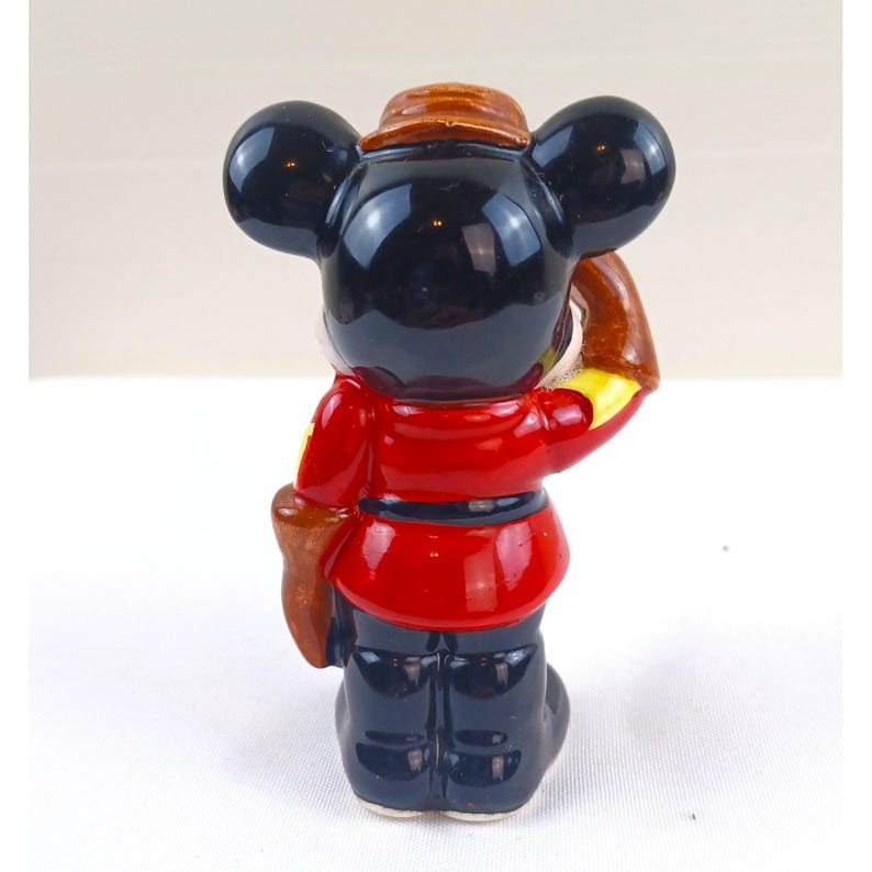 May include: A ceramic figurine of a cartoon mouse wearing a red jacket, dark blue pants, and a brown hat. The mouse has large black ears and is holding a brown object. The figurine is standing on a white surface.