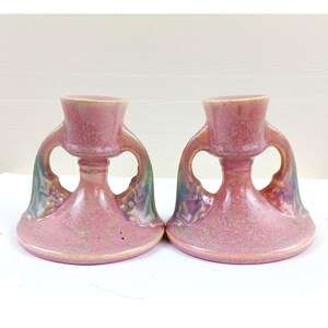 Puede incluir: Dos candeleros de cerámica rosa a juego. Cada uno tiene una parte superior en forma de copa, dos asas curvas y una base acampanada. Las asas tienen un diseño decorativo con toques de verde y morado. Los candeleros tienen un acabado brillante.