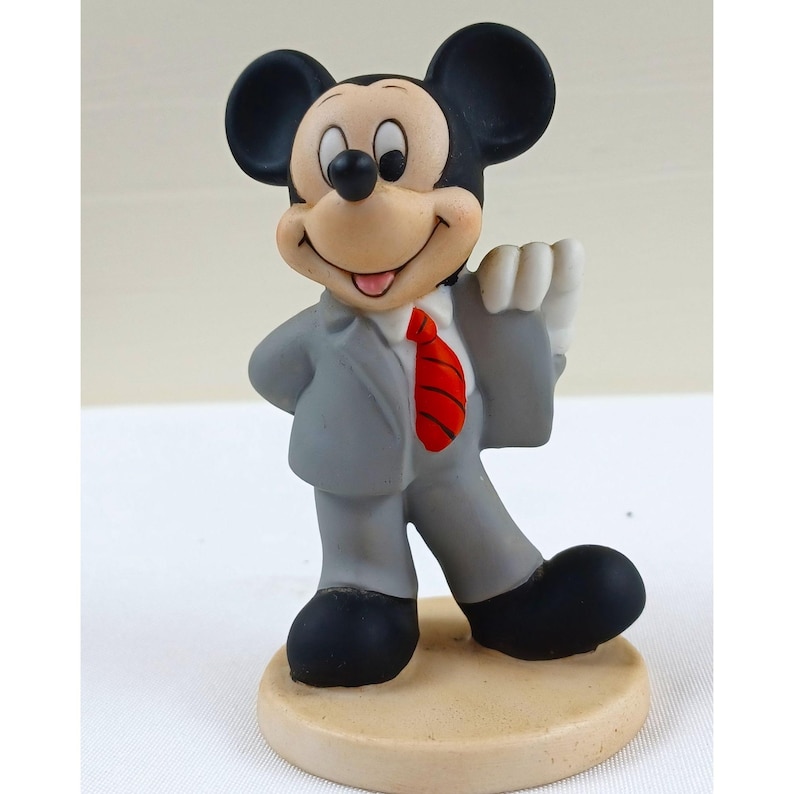 Pode incluir: Uma estatueta do Mickey Mouse em um terno cinza com uma gravata vermelha, sapatos e orelhas pretas. A figura est&aacute; em uma base bege e tem uma express&atilde;o amig&aacute;vel. Um item de colecionador.
