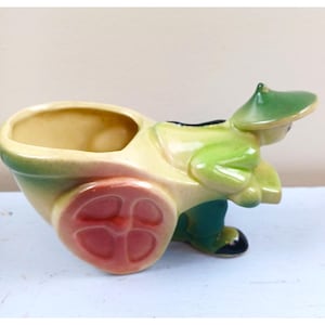May include: A vintage ceramic planter in the shape of a man pushing a wheelbarrow. The planter is painted in shades of yellow, green, and pink. The wheel has a pink center. The man is wearing a green hat and pants.