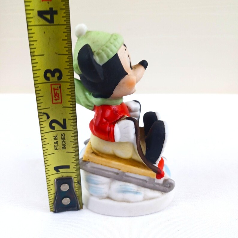 May include: A ceramic figurine of Mickey Mouse on a sled. Mickey is wearing a red jacket, green hat, and scarf. The figurine is approximately 7.6 cm tall.