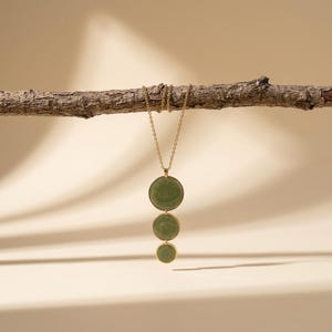 May include: A gold-toned necklace featuring three circular, sage green stone pendants arranged vertically in descending order of size. The pendants are encased in thin gold metal rims and suspended from a delicate gold-toned metal chain.