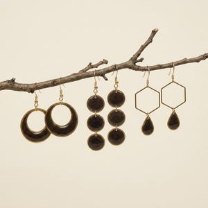 May include: Three pairs of gold-toned earrings hanging from a tree branch. The first pair features large circular hoops, the second pair consists of three stacked dark circular discs, and the third pair displays a hexagonal frame with a teardrop charm.