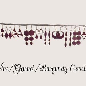 May include: A collection of wine-colored earrings in various shapes, including teardrops, diamonds, and geometric designs, hang from a thin branch. Some earrings feature gold-colored accents. The text "Wine / Garnet / Burgundy Earrings" is at the bottom.