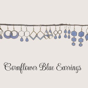 May include: A collection of cornflower blue earrings hangs from a branch. The earrings feature various geometric shapes, including teardrops, circles, and squares, with gold-colored metal accents. The text "Cornflower Blue Earrings" is displayed in elegant script below.