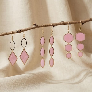 May include: Three pairs of gold-toned dangle earrings hanging from a branch. The first pair features a black oval hoop with a pink diamond-shaped pendant. The second pair has three vertical pink marquise shapes. The third pair has three pink hexagons.