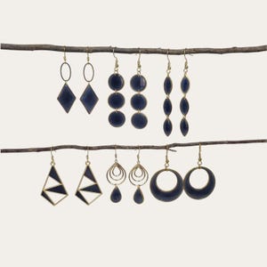 May include: Six pairs of gold-toned earrings with dark blue enamel accents, displayed on two horizontal branches. The collection includes diamond, circular, marquise, geometric triangle, teardrop, and crescent moon shapes with hook-style backings.