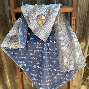 Puede incluir: Una manta para bebé suave, gris y azul con un parche con temática desértica. La manta presenta un lado azul marino con flechas blancas y un lado gris texturizado. El parche representa cactus, una luna y un sol.
