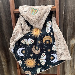 Puede incluir: Una manta con capucha para bebé con un diseño celestial. La manta presenta un fondo negro con soles, lunas, estrellas y polillas. La capucha y el ribete son de un material de felpa, color canela. Un parche de mariposa bordado está en la capucha.