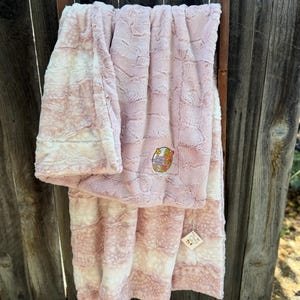 Könnte beinhalten: Eine weiche, rosa-weiße Babydecke mit strukturierter Oberfläche ist über eine Holzleiter drapiert. Die Decke hat einen farbenfrohen gestickten Aufnäher und ein kleines Etikett. Die Decke ist aus einem Plüschmaterial.