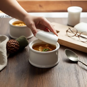 May include: White ceramic soup bowls with lids, filled with soup and garnished with herbs. One bowl has its lid slightly ajar, revealing the contents. The bowls are on a wooden table, with a spoon and glasses in the background.