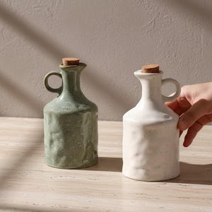 Puede incluir: Dos botellas de cerámica para aceite de oliva con tapones de corcho. Una es verde apagado, la otra blanquecina. Ambas tienen un asa pequeña y una superficie texturizada. Las botellas se exhiben sobre una superficie clara.