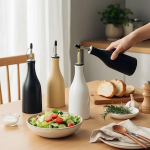May include: A collection of ceramic oil and vinegar bottles in black, beige, and white, with stainless steel pour spouts. One bottle is being used to pour oil. A bowl of salad and bread slices are also on the table.