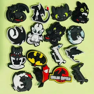 May include: A selection of black and white cartoon shoe charms, featuring dragon and bat designs. Some charms include text. The charms are displayed against a light green background.