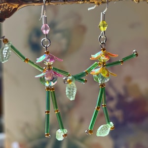 Puede incluir: Un par de pendientes colgantes hechos a mano con diseños caprichosos de hadas. Los pendientes tienen cuerpos de cuentas verdes, cabezas iridiscentes en forma de flor y detalles de hojas. Los pendientes cuelgan de ganchos plateados y están adornados con cuentas rosas y amarillas.