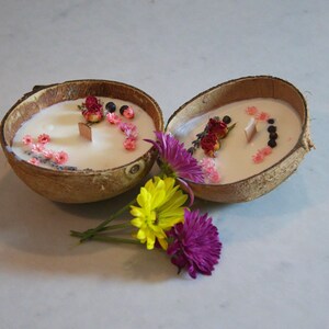 May include: Two coconut shell candles with white wax, decorated with dried flowers and wooden wicks. The candles are surrounded by yellow and purple flowers, set on a white surface.